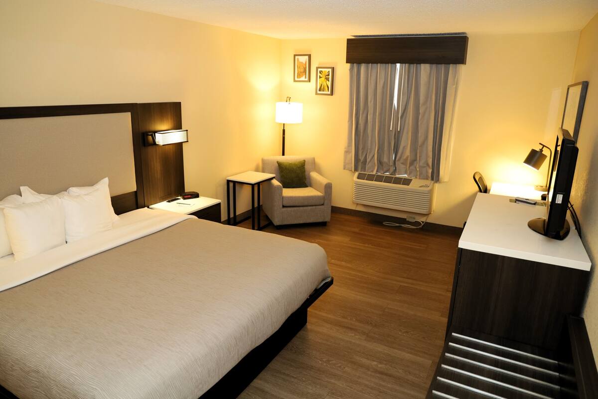 A hotel room with a large bed, a desk, a chair, a lamp, an air conditioning unit, and a window with curtains.