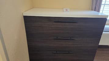 A dark wooden dresser with a white top and three drawers, located in a corner of a room with a beige wall and a window with blinds.