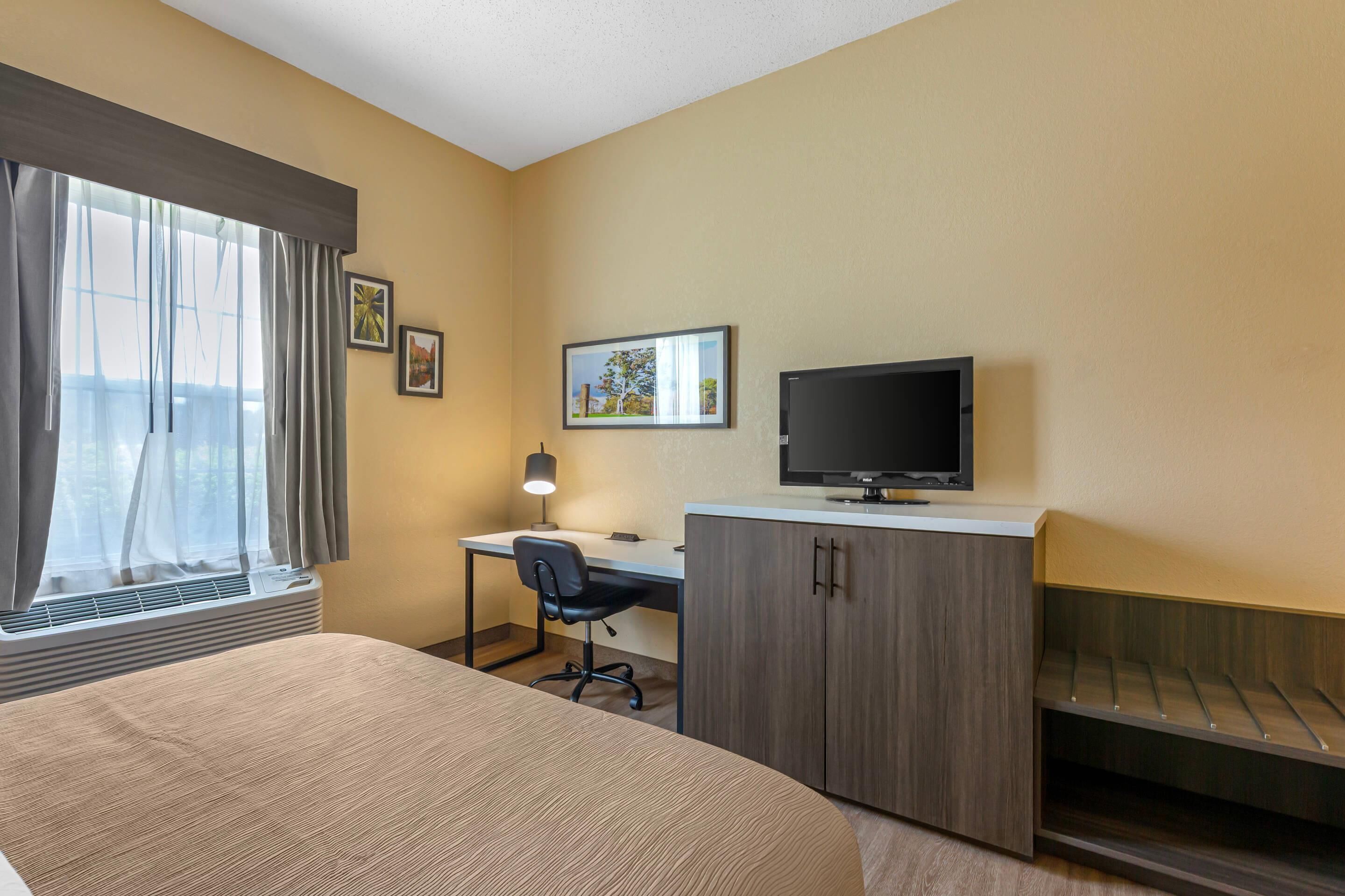 A hotel room with a bed, a desk with a chair, a television, and a window with curtains.