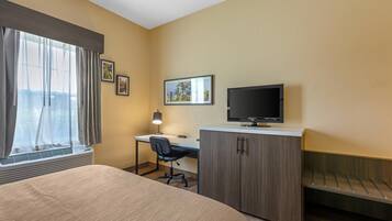 Quarto de hotel com cama, escrivaninha com cadeira, televisão e janela com cortinas.