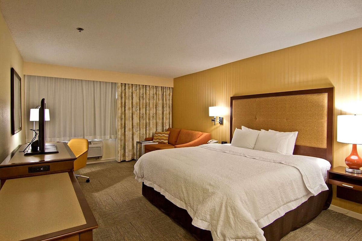 A hotel room with a large bed, a desk with a lamp, a chair, and a window with curtains.