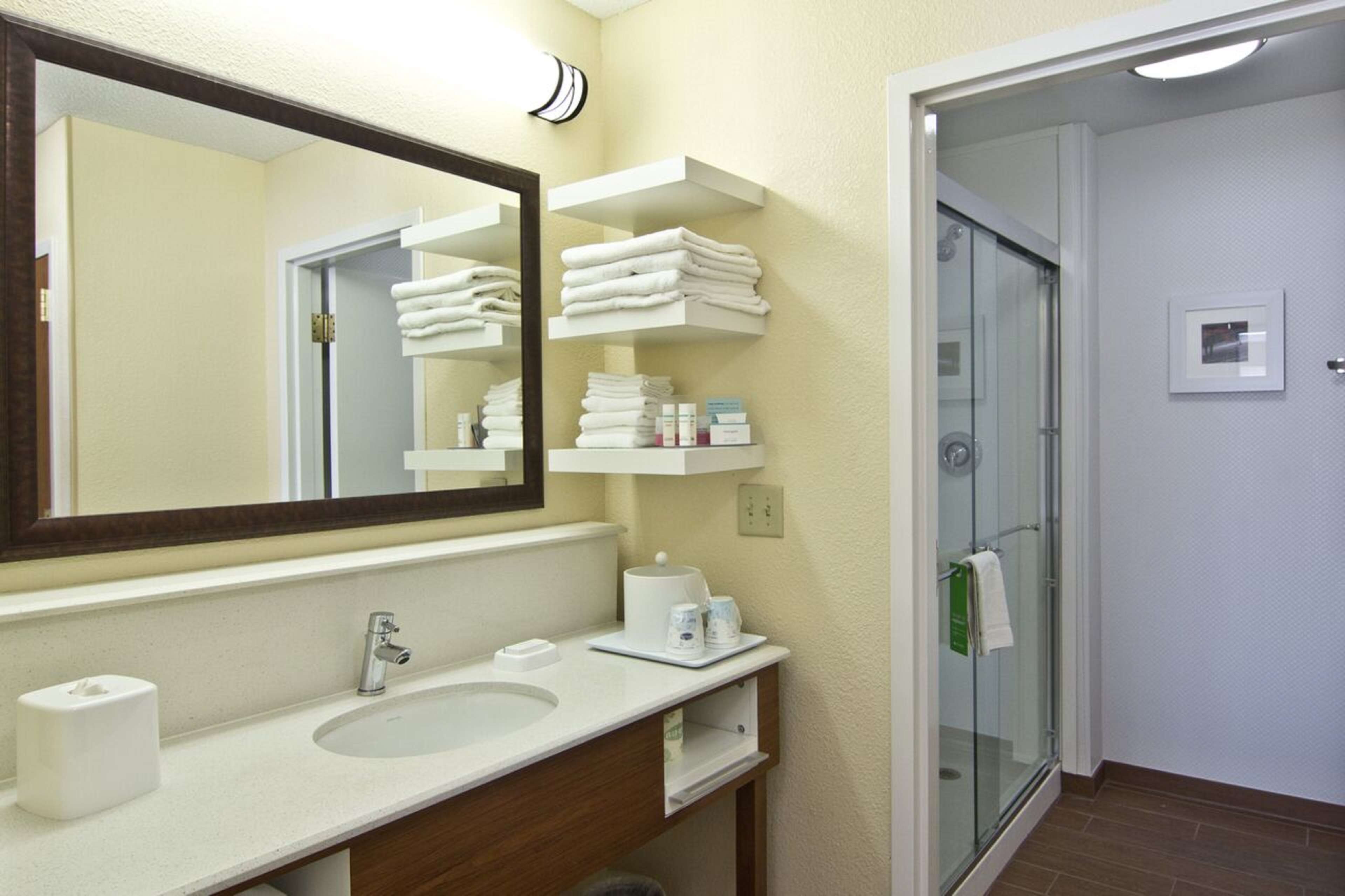A hotel bathroom with a large mirror, a sink, a glass shower enclosure, and shelves with towels and toiletries.