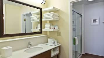 A hotel bathroom with a large mirror, a sink, a glass shower enclosure, and shelves with towels and toiletries.