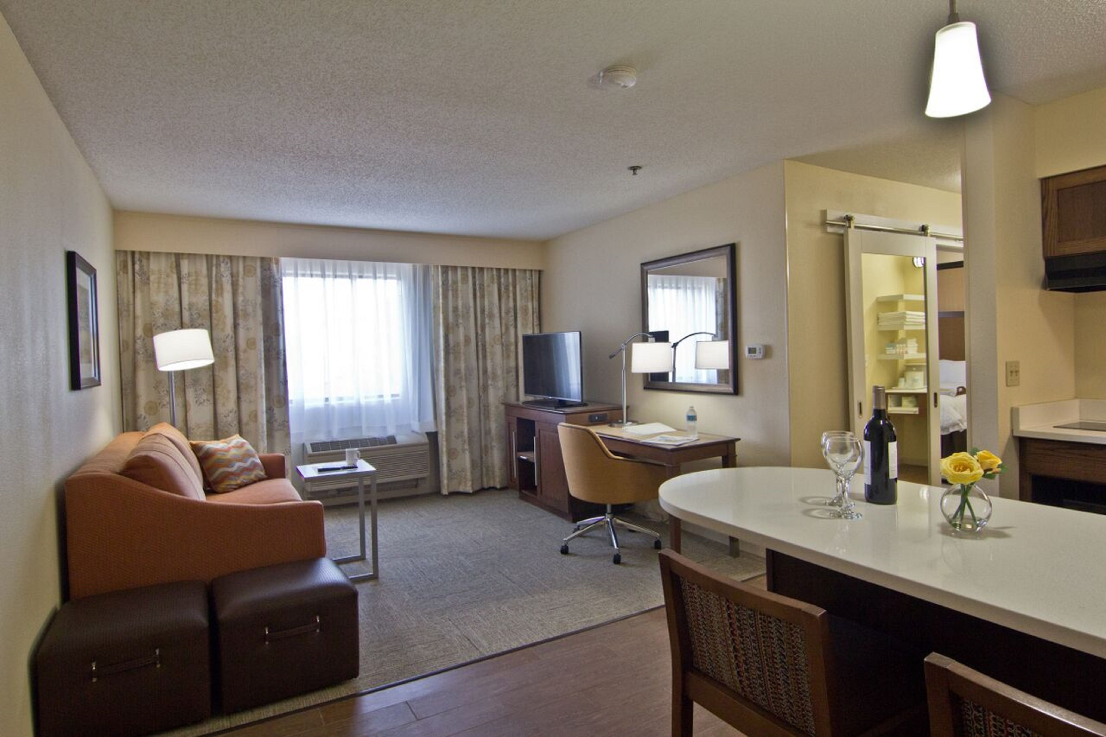 A hotel room with a sofa, a desk, a television, a dining table, and a kitchen area.