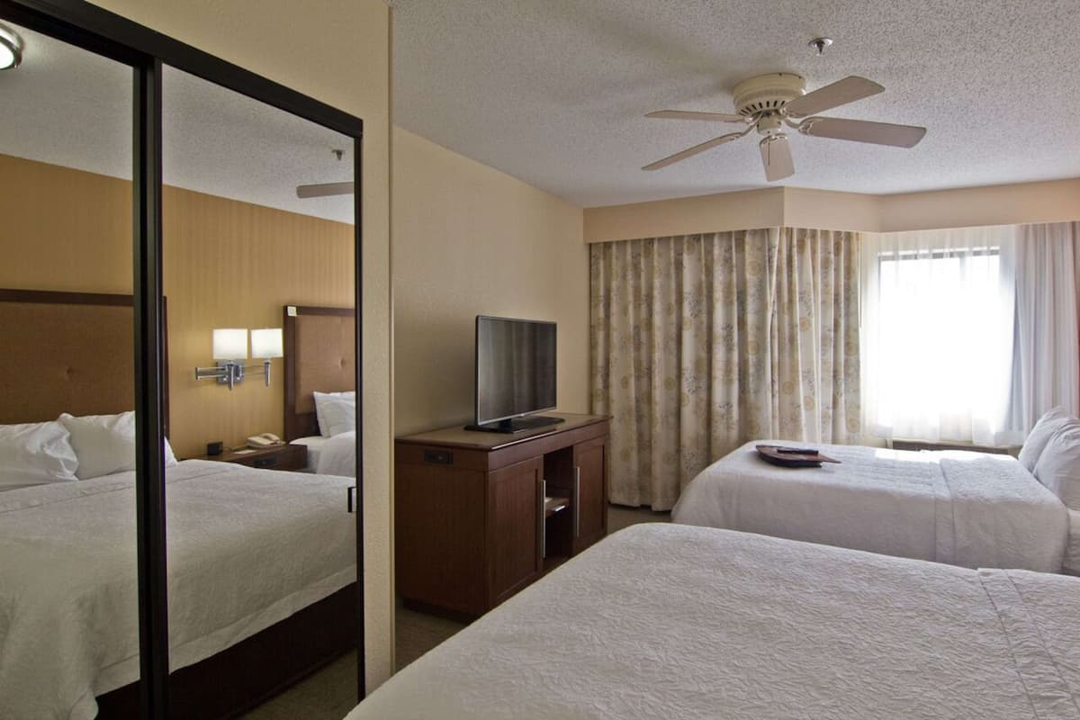A hotel room with two beds, a television on a wooden cabinet, a ceiling fan, and a large window with curtains.