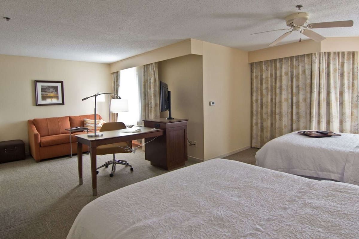 A hotel room with two beds, a desk with a chair, a sofa, a ceiling fan, and a large window with curtains.