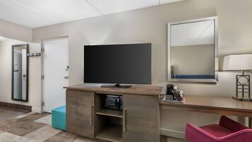 A hotel room with a flat-screen TV, a desk with a coffee maker, a mirror, and a pink chair.