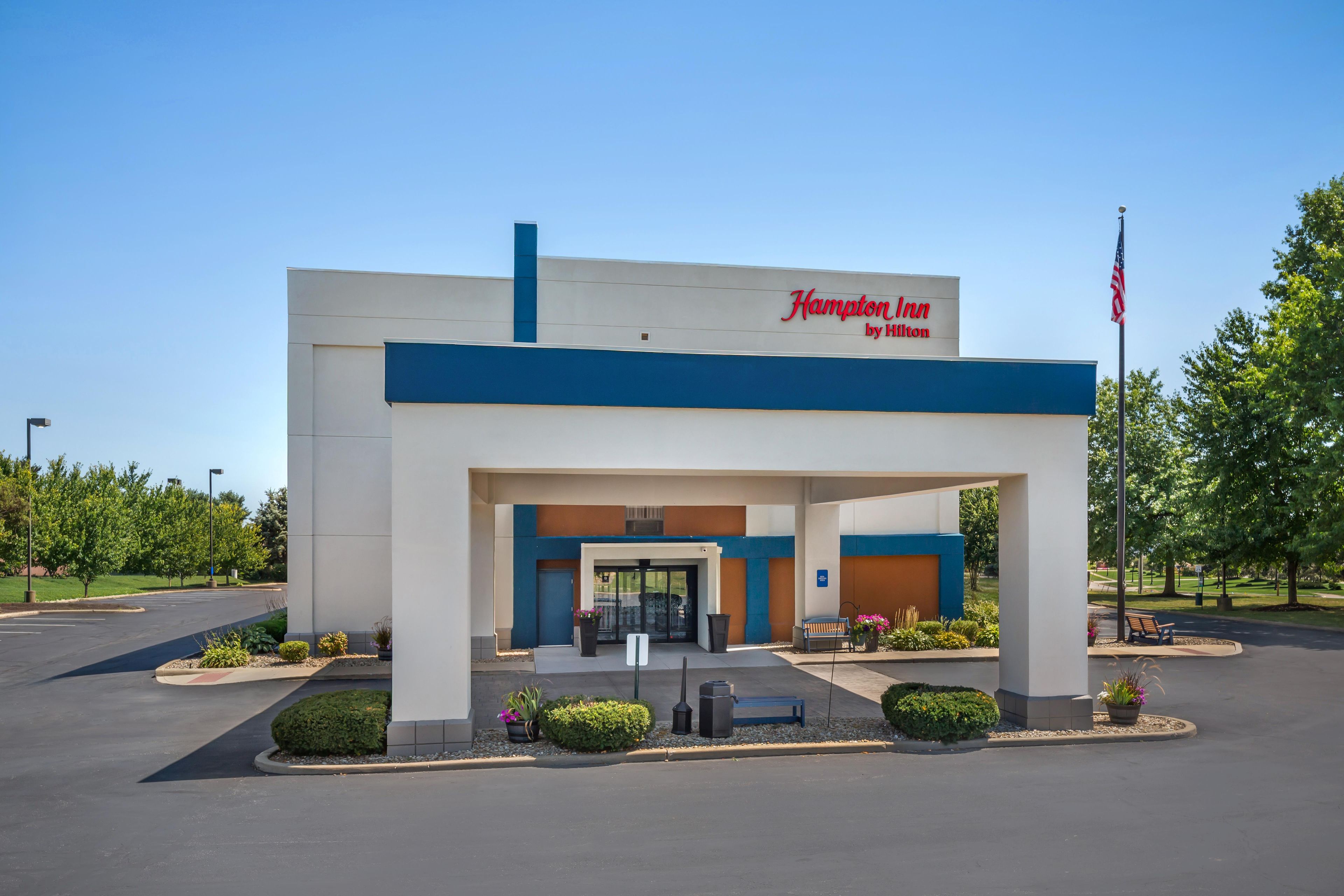 Un Hampton Inn by Hilton con entrada despejada, bandera estadounidense y jardines bien cuidados.
