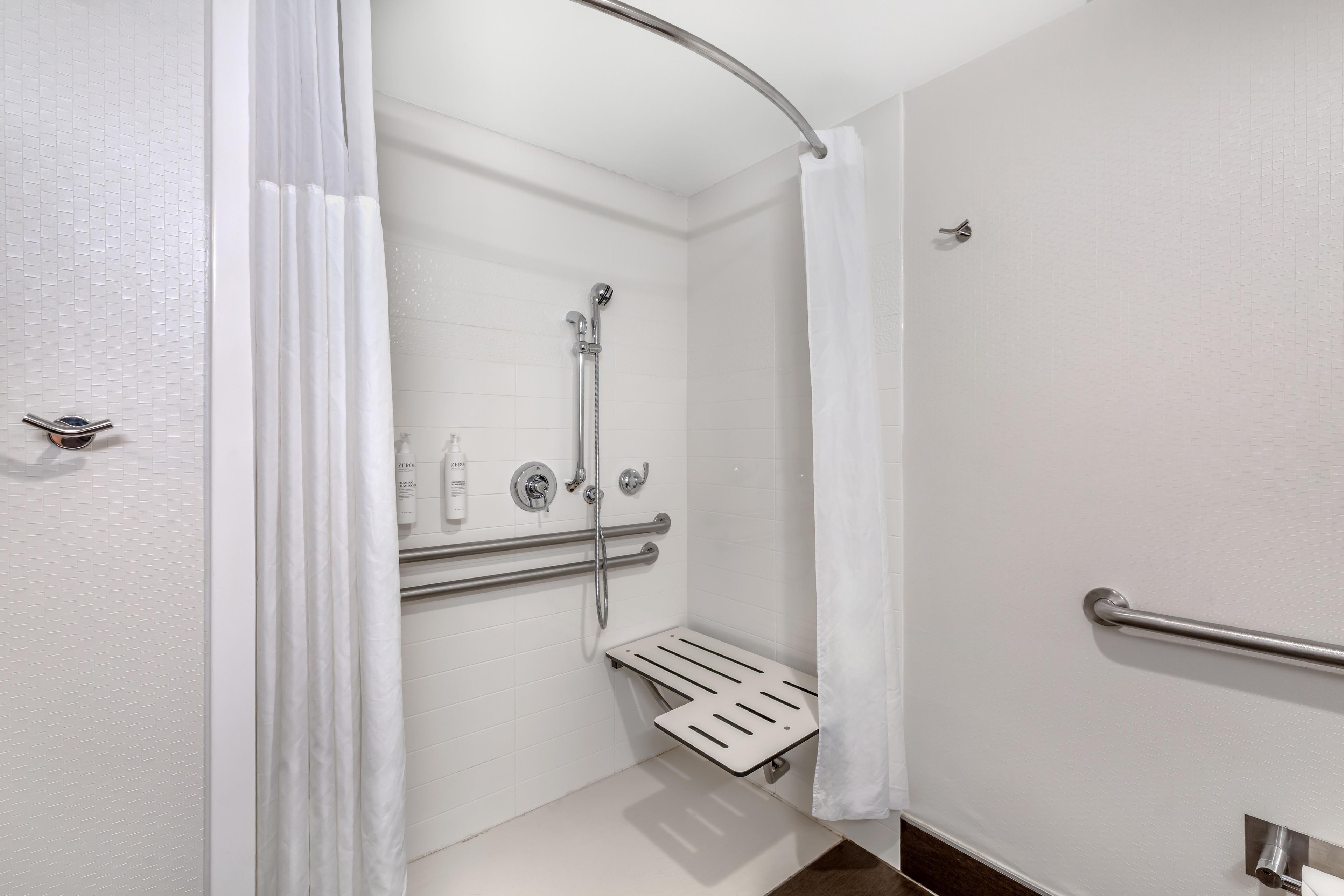 Un baño moderno con paredes de azulejos blancos, una ducha con cabezal de ducha manual, una barra de apoyo y un perchero blanco.