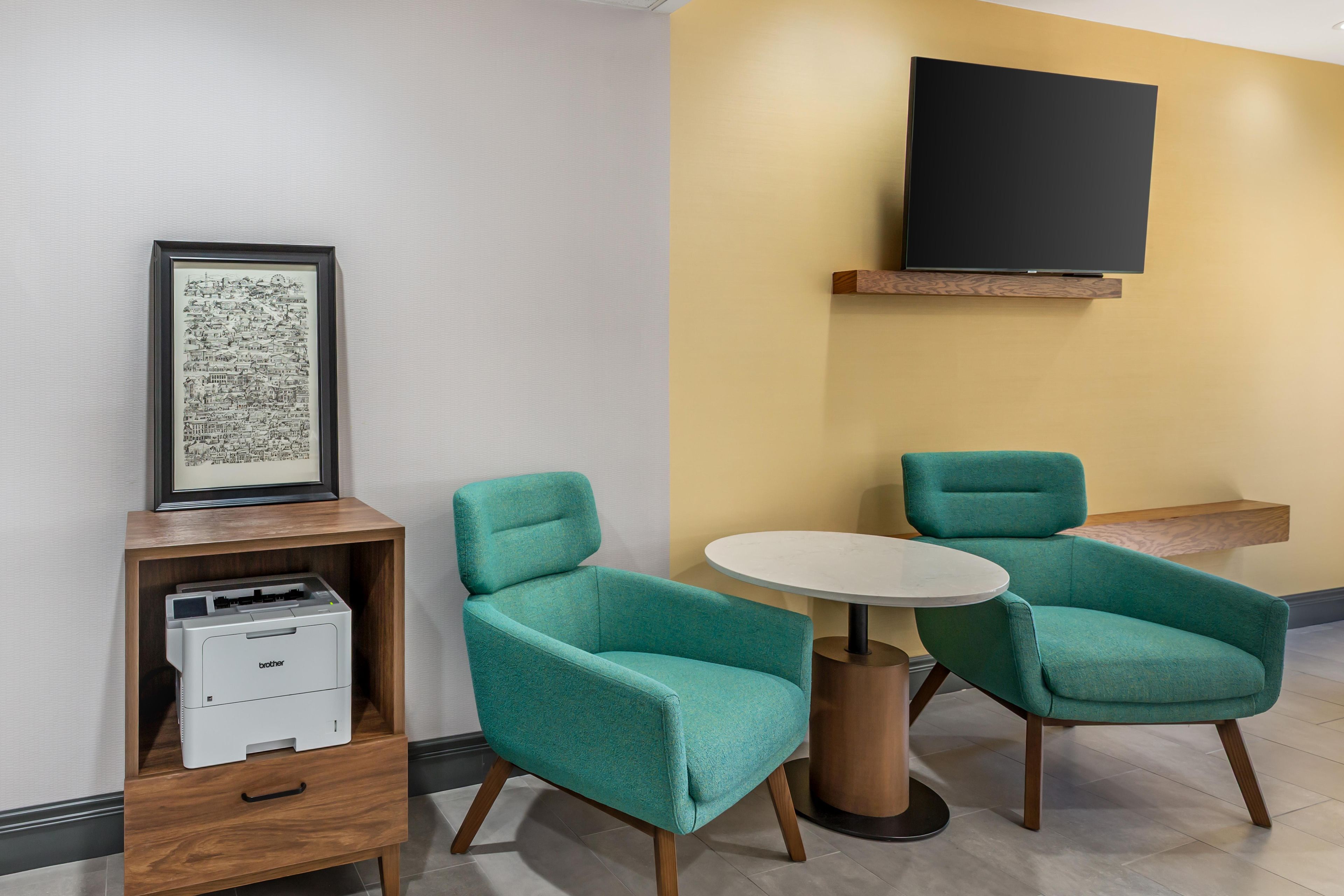A room with teal chairs, a white table, a framed cityscape artwork, a printer, and a mounted TV on the wall.