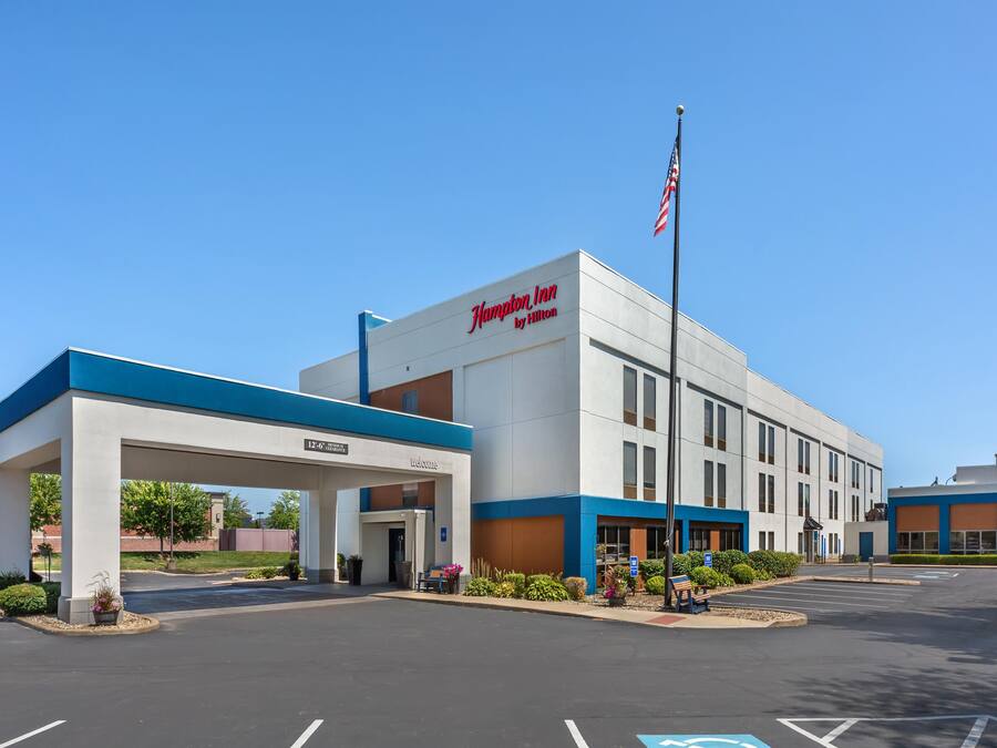A Hampton Inn by Hilton hotel with a covered entrance, parking lot, and American flag.