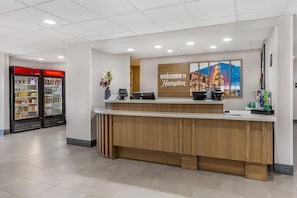 Una mesa de recepción con un cartel que dice 'Welcome to Hampton', una máquina de café, un jarrón con flores y una obra de arte colgada en la pared.