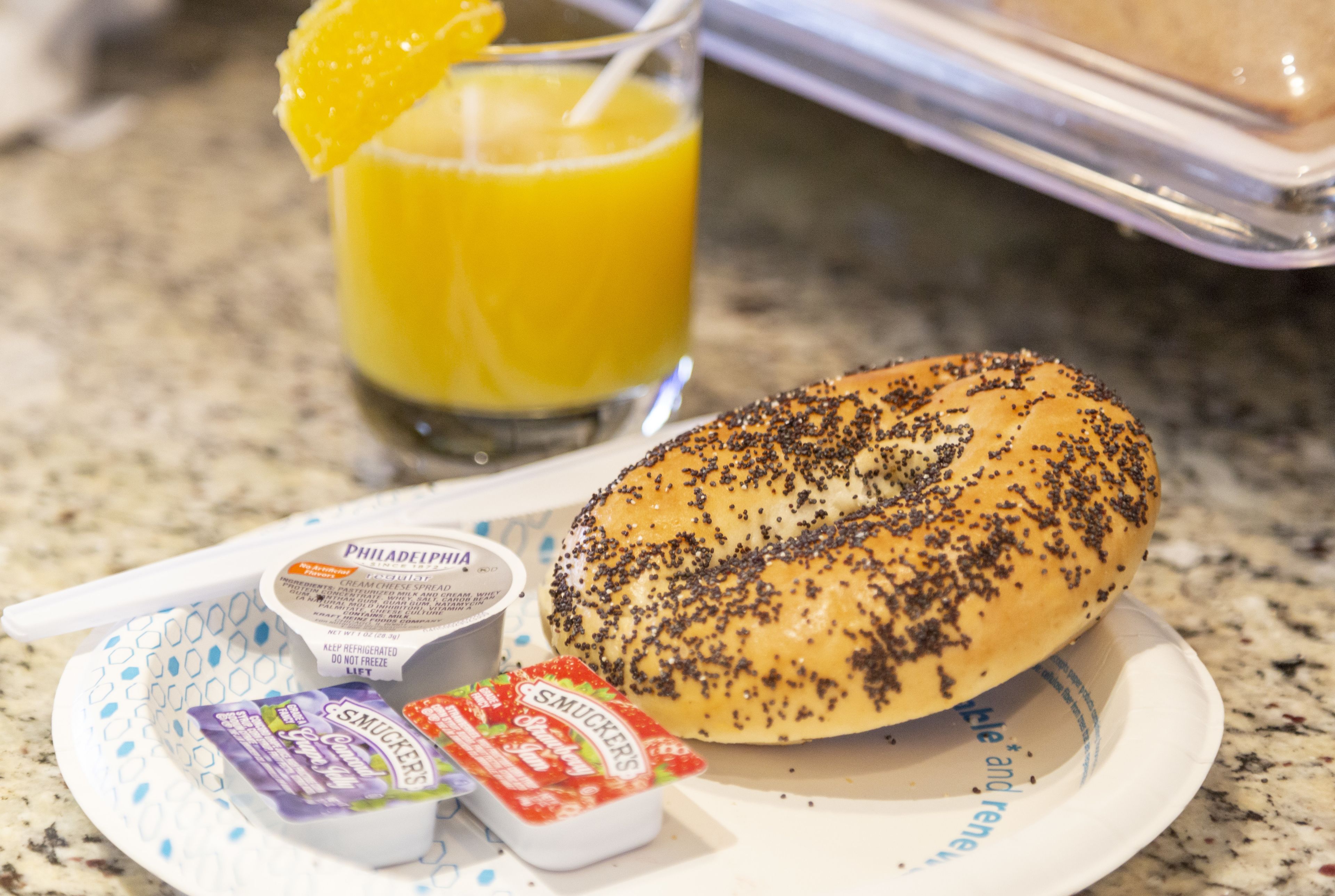 Een ontbijfwagen met een bagel belegd met papzaadjes, een glas sinaasappelsap en twee bakjes roomkaas en jam.