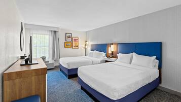 Ein Hotelzimmer mit zwei Betten, einem Schreibtisch, einem Stuhl, einem Fenster mit Vorhängen und Wandbildern.