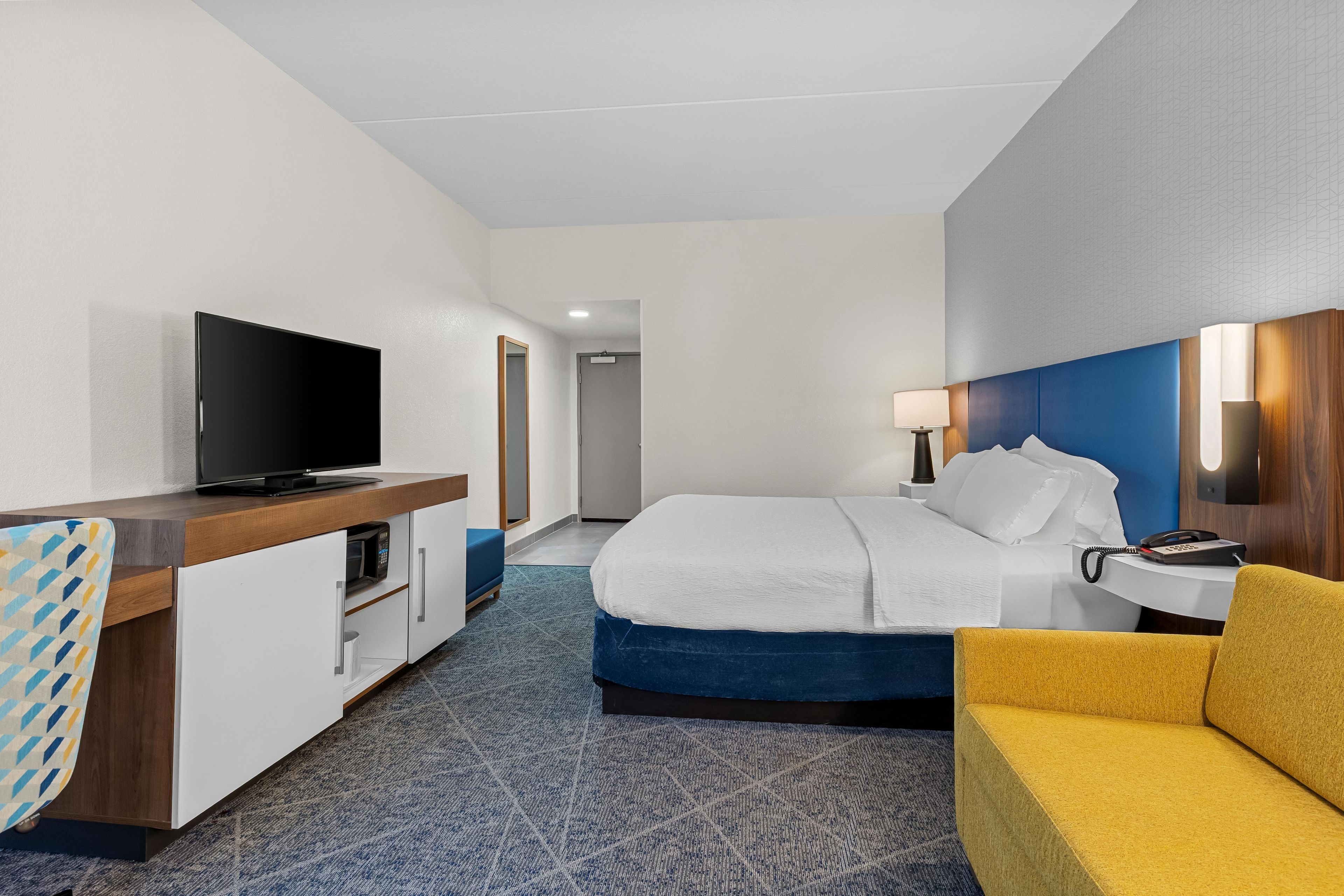 A hotel room with a bed, a desk with a television, a bedside table with a lamp, and a sofa.