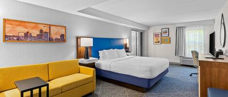 A hotel room with a bed, a yellow sofa, a desk, a chair, and a cityscape painting on the wall.