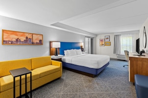 A hotel room with a bed, a yellow sofa, a desk, a chair, and a cityscape painting on the wall.