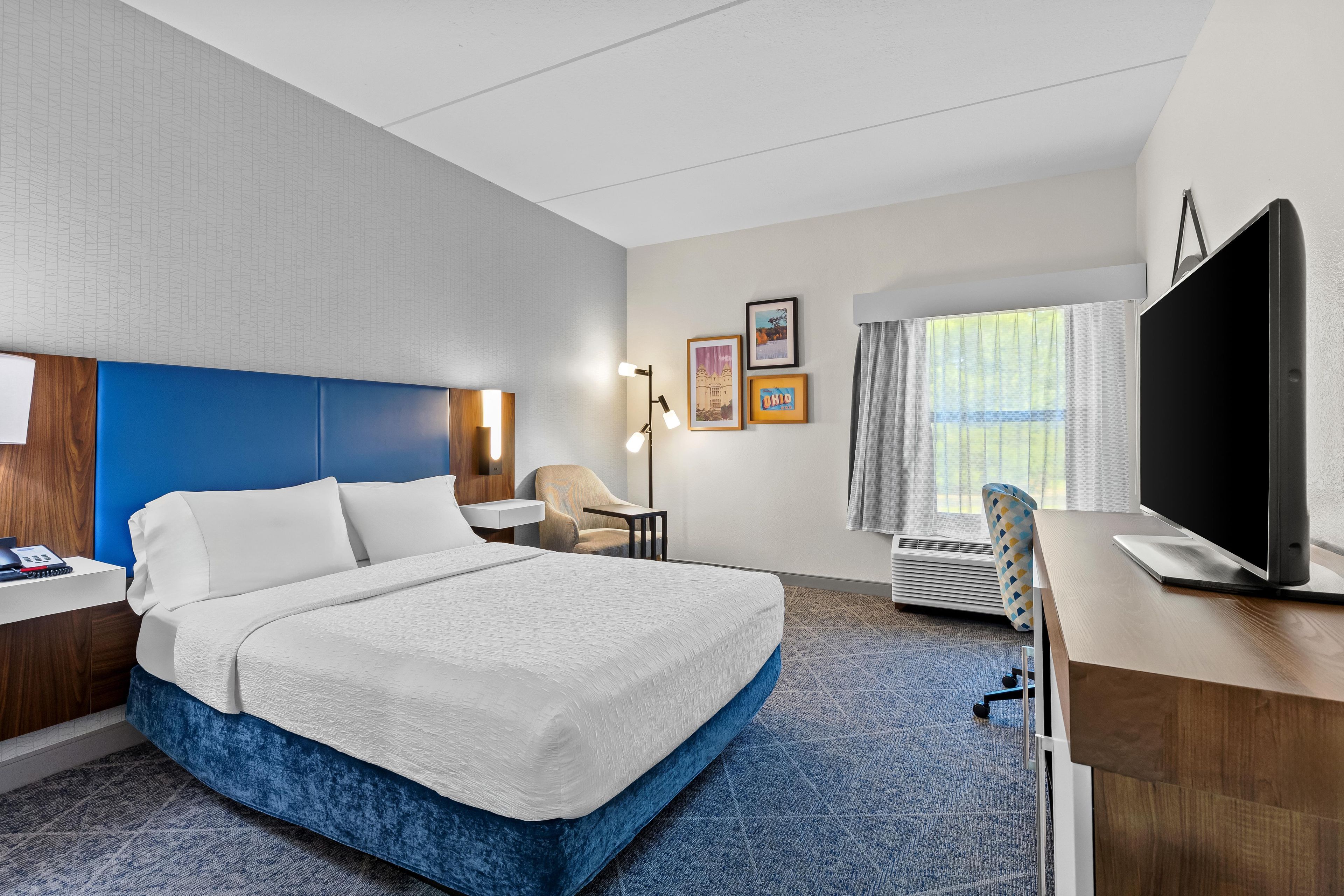 Quarto de hotel com cama, escrivaninha, cadeira e televisão.