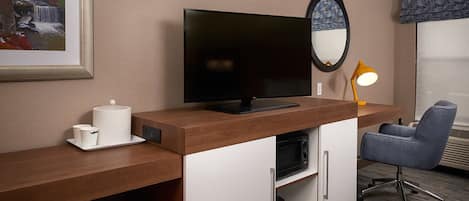 A hotel room with a wooden desk, a flat-screen TV, a microwave, a red chair, a small table with a white mug, and a framed picture of a waterfall.