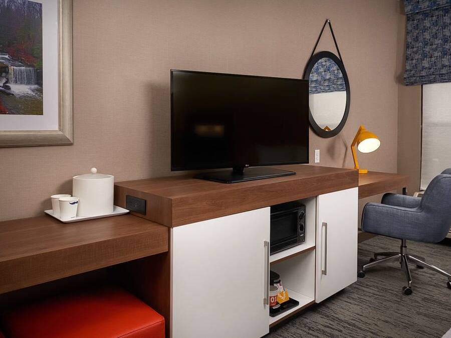 Habitación de hotel con escritorio de madera, televisor de pantalla plana, microondas, una silla roja, una mesita con una taza blanca y un cuadro con una cascada.