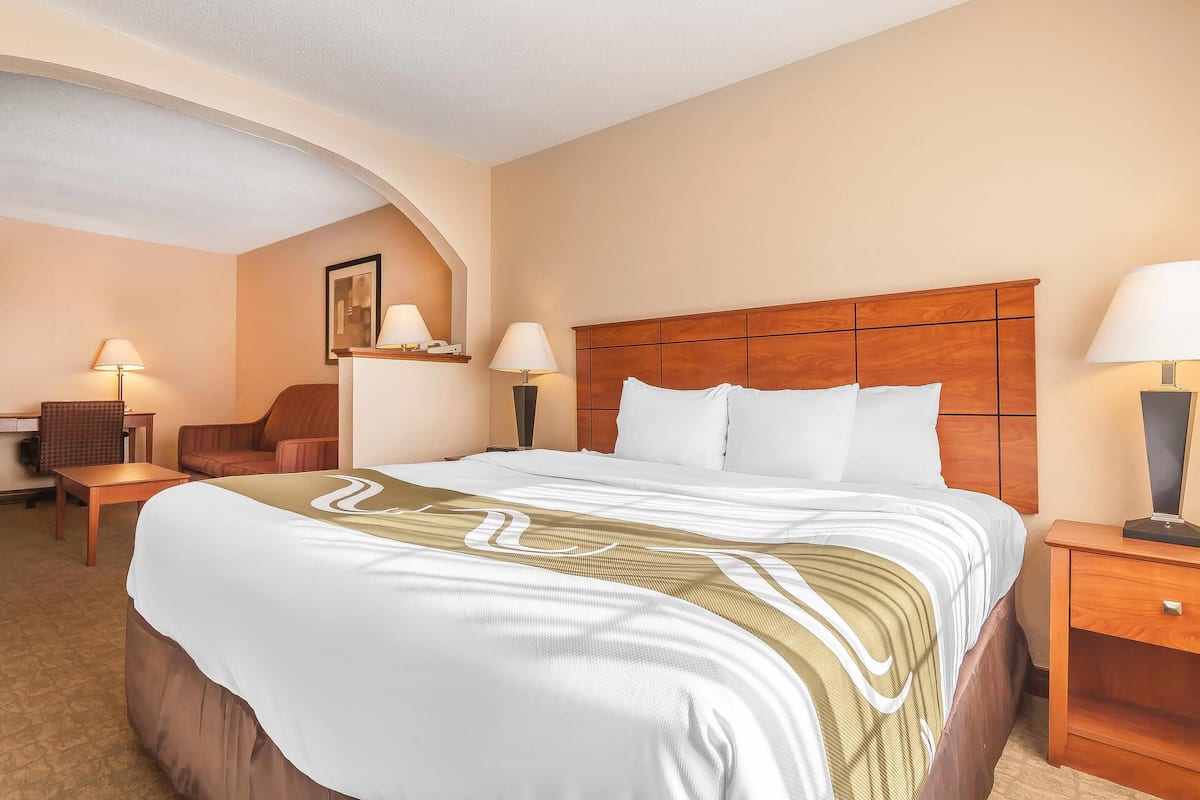 A hotel room with a large bed, two bedside tables with lamps, a wooden headboard, a desk with a chair, and a small seating area with a chair and table.