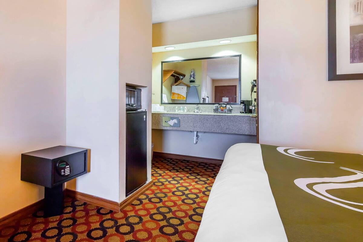 A hotel room with a bed, a small safe, a mirror, a sink, and a patterned carpet.