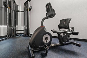 Una bicicleta estática, una máquina de pesas y un estante fijado a la pared en un gimnasio.