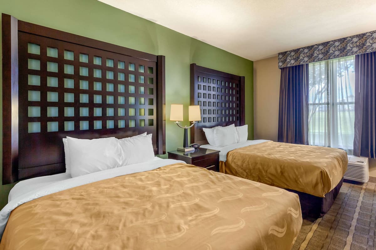 A hotel room with two beds, a headboard with a grid pattern, a nightstand with a lamp, and a window with curtains.