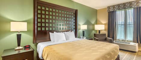 A hotel room with a large bed, two bedside tables with lamps, a headboard with a grid pattern, a window with curtains, and a wall-mounted air conditioner.