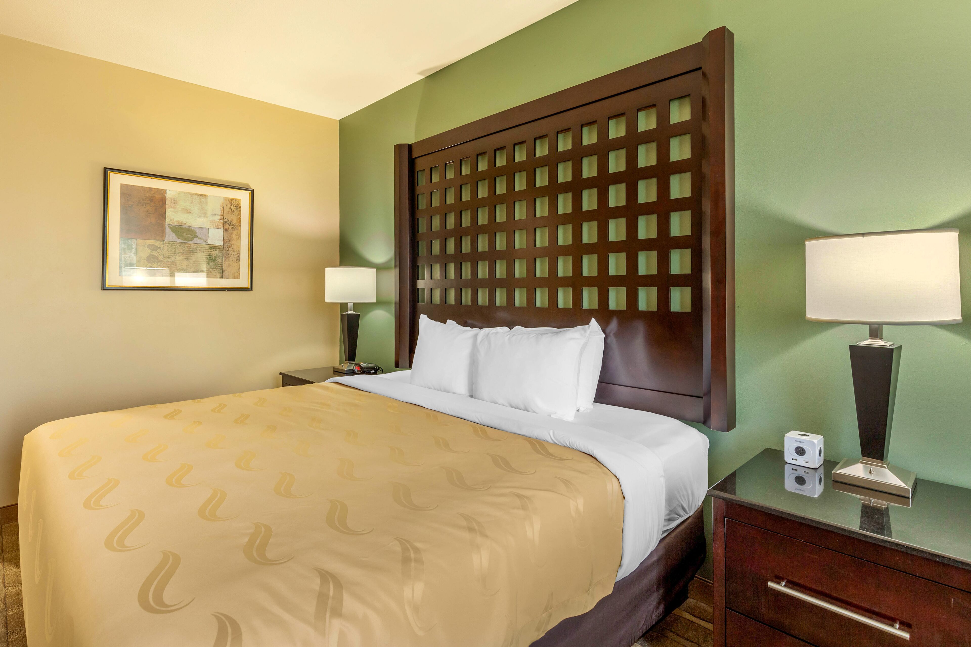 A hotel room with a large bed, a wooden headboard, two bedside tables with lamps, a framed picture on the wall, and a green accent wall.