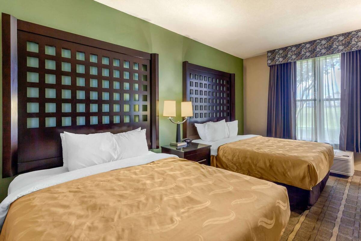 A hotel room with two beds, a nightstand, a lamp, and a window with curtains.