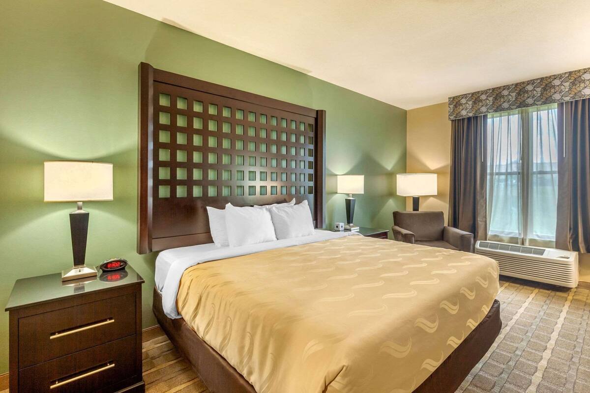 A hotel room with a large bed, two bedside tables, a nightstand, a chair, a window with curtains, and an air conditioning unit.