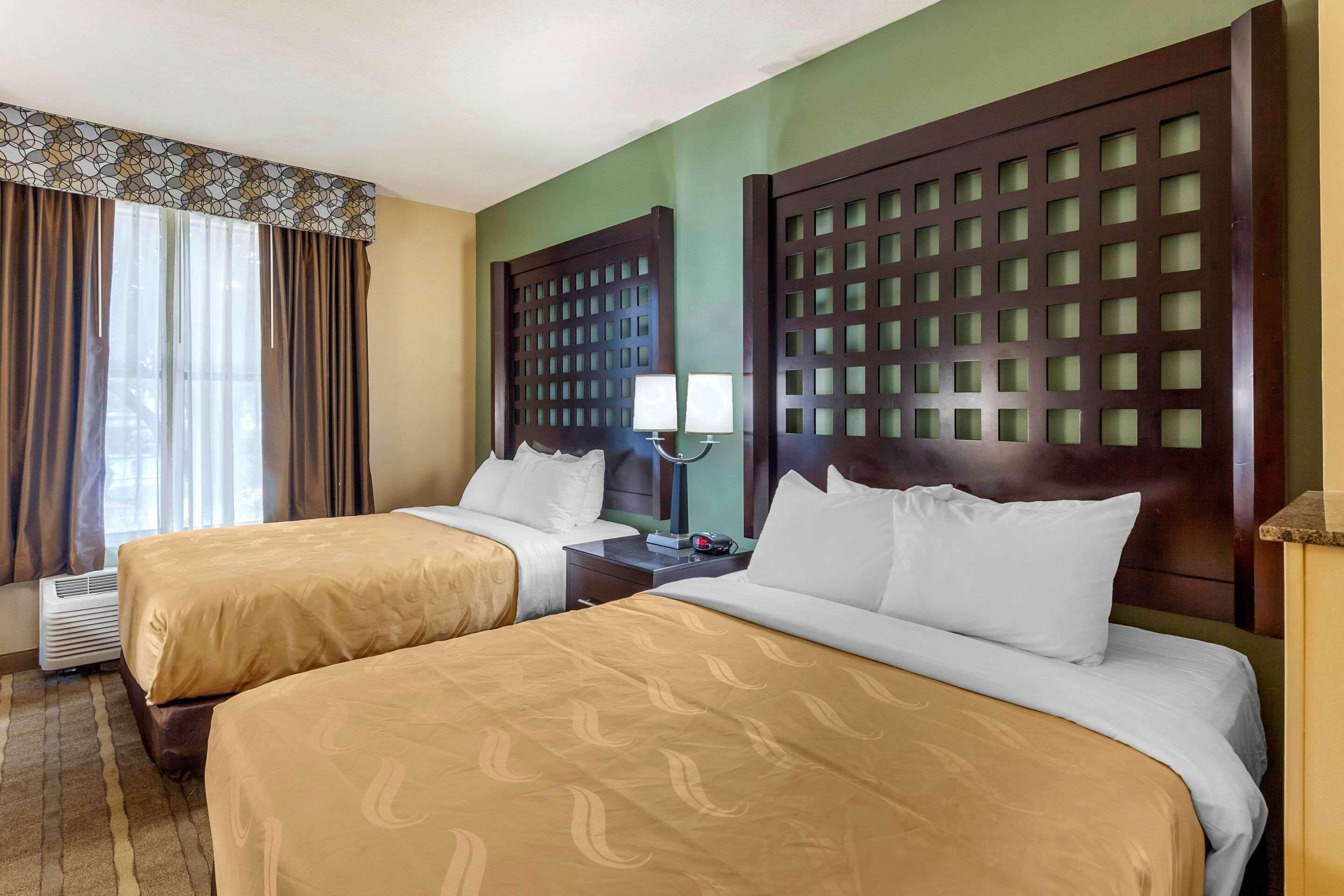 A hotel room with two beds, a nightstand with a lamp, a window with curtains, and a wall-mounted air conditioner.