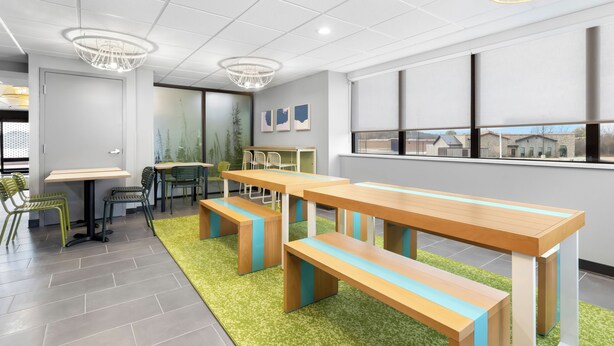 Un espace de restauration moderne avec des tables et des bancs en bois, des chaises vertes et une vue sur les bâtiments à travers de grandes fenêtres.