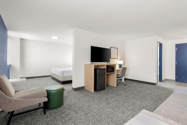 A hotel room with a bed, a desk with a computer, a chair, a trash bin, and a TV on the wall.