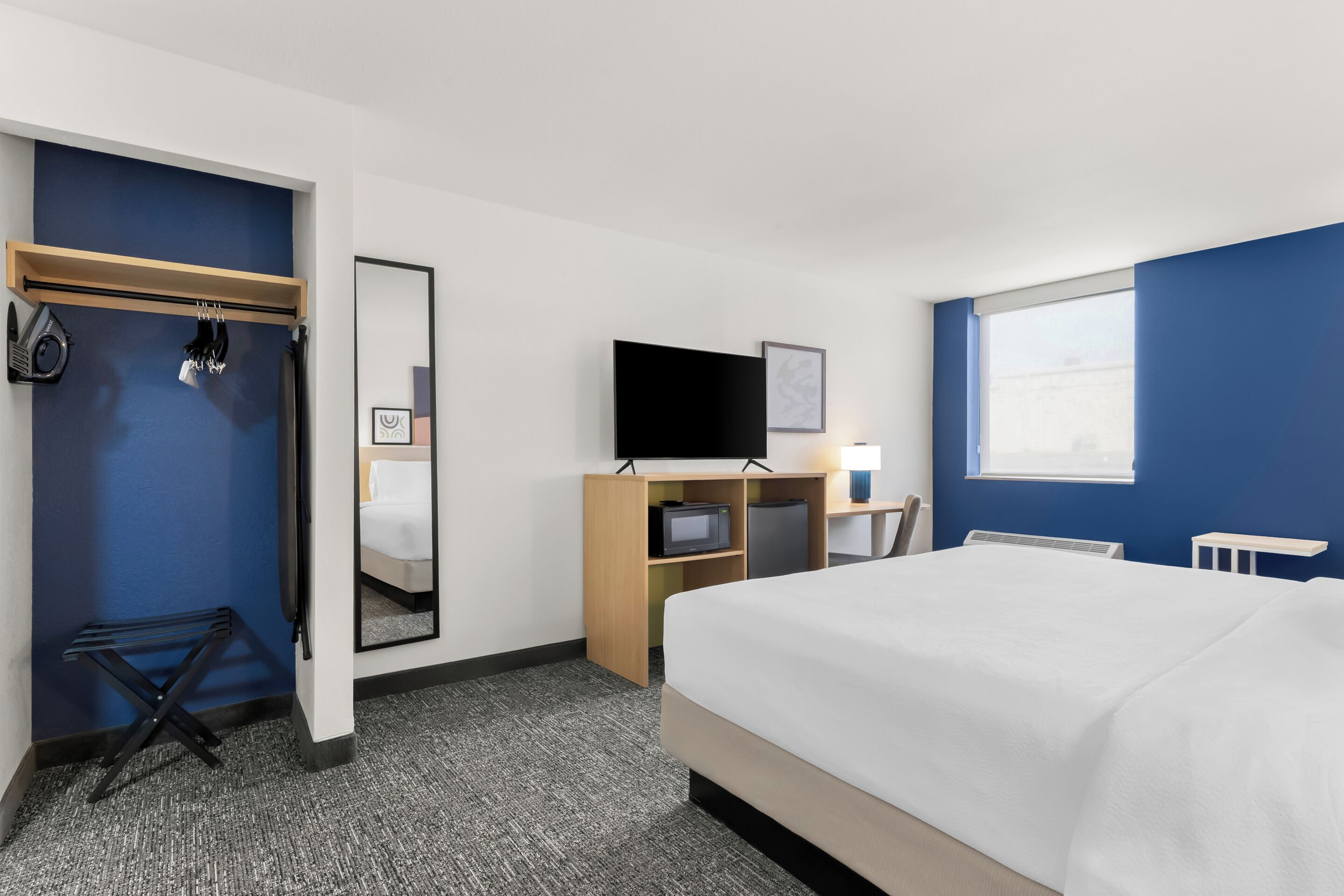 A hotel room with a bed, a desk with a TV, a mirror, and a window.