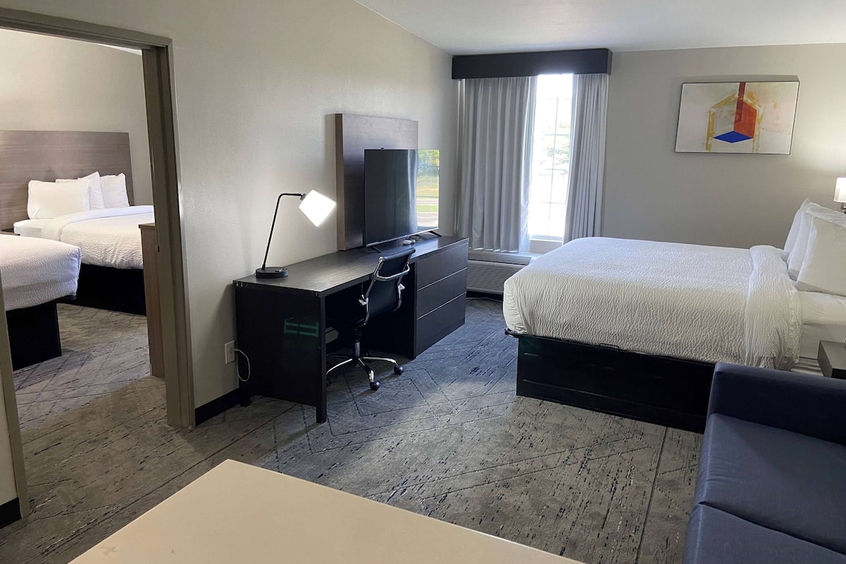 A hotel room with two beds, a desk with a computer, a TV, and a large window with curtains.