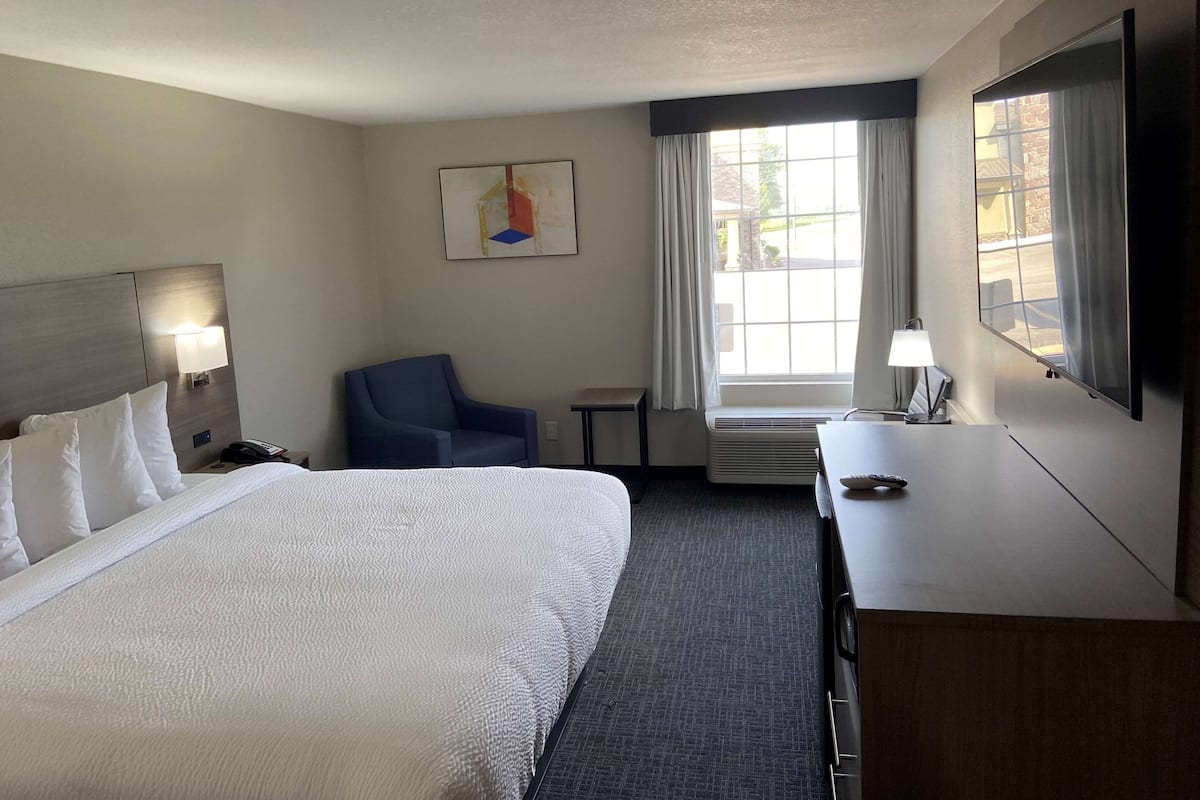 A hotel room with a large bed, a desk, a chair, a small table, a lamp, a painting, and a window with curtains.