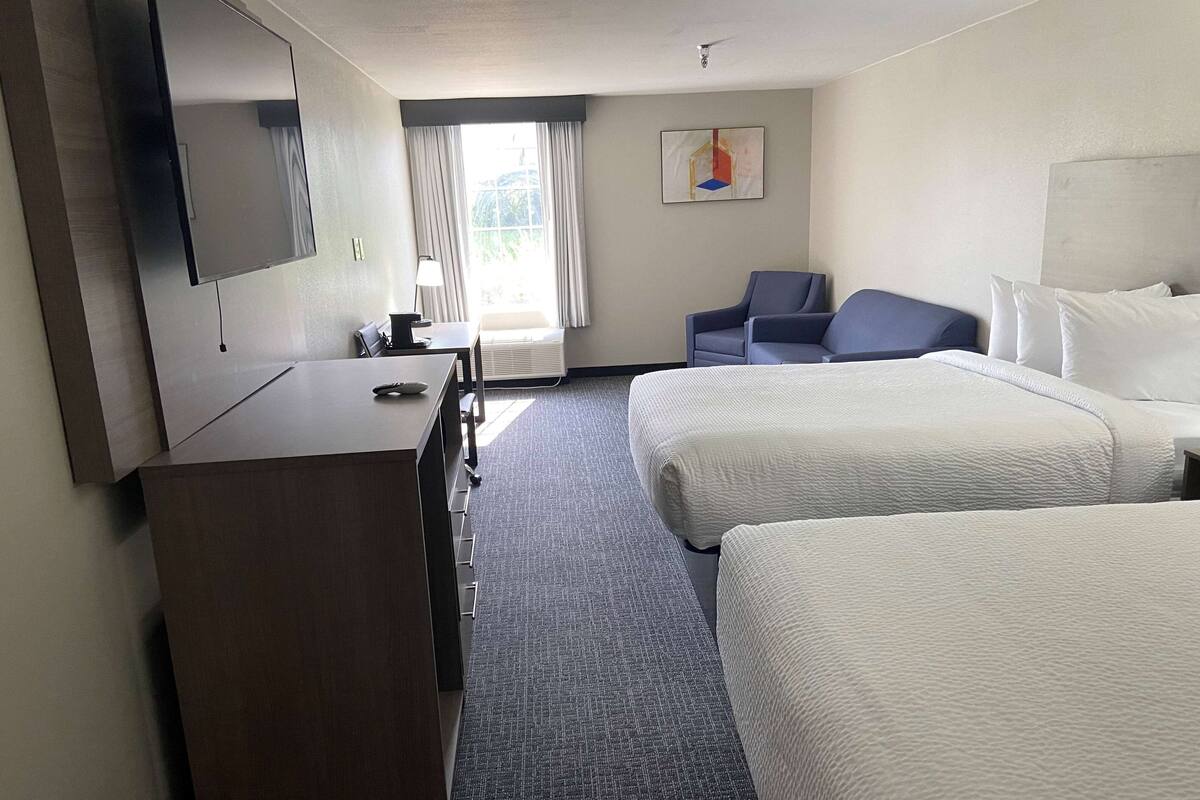 A hotel room with two beds, a desk, a chair, a flat-screen TV, and a window with curtains.