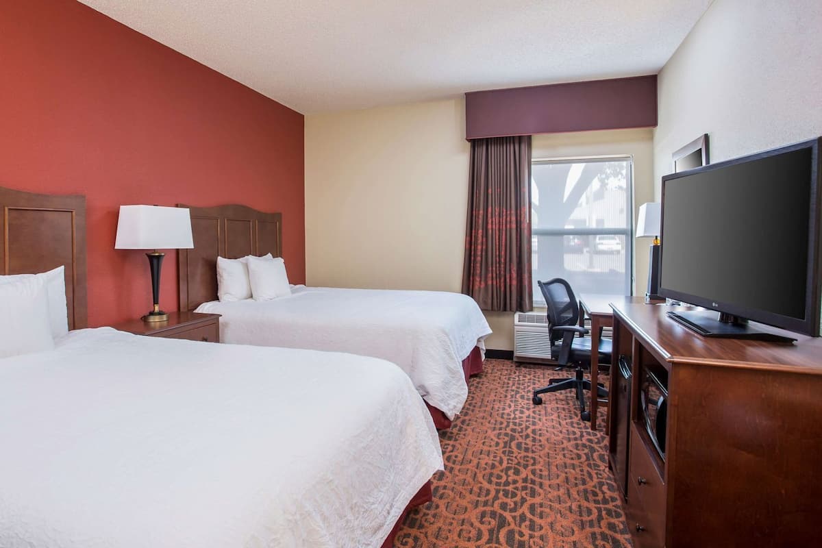 A hotel room with two beds, a desk with a TV, a chair, and a window with curtains.