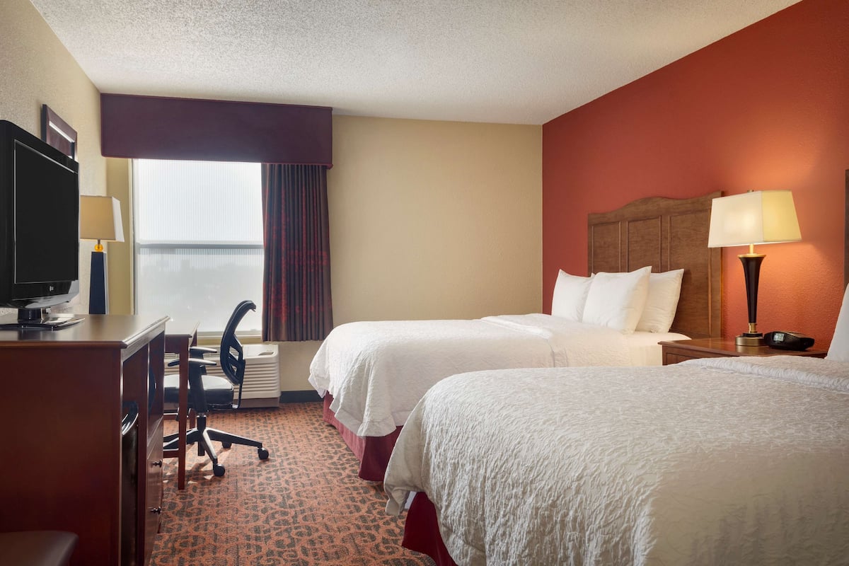 A hotel room with two beds, a desk with a computer, a chair, a TV, and a window with curtains.