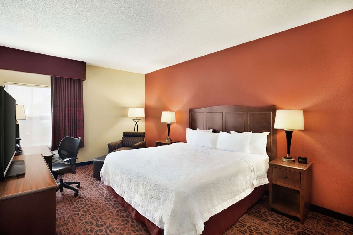A hotel room with a large bed, two bedside tables with lamps, a desk with a chair, and a window with curtains.