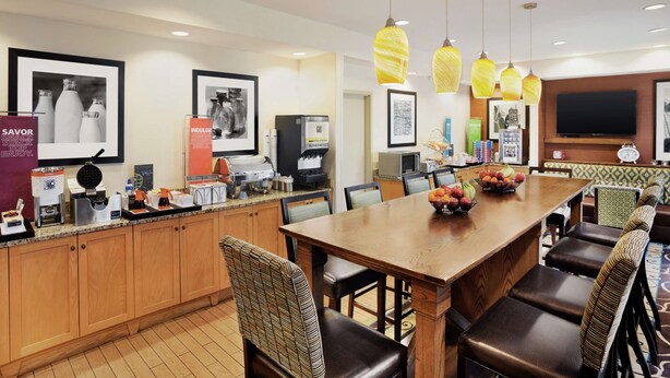 A hotel dining area with a wooden table, chairs, and a fruit bowl. There are hanging lights, framed pictures, and a television on the wall.