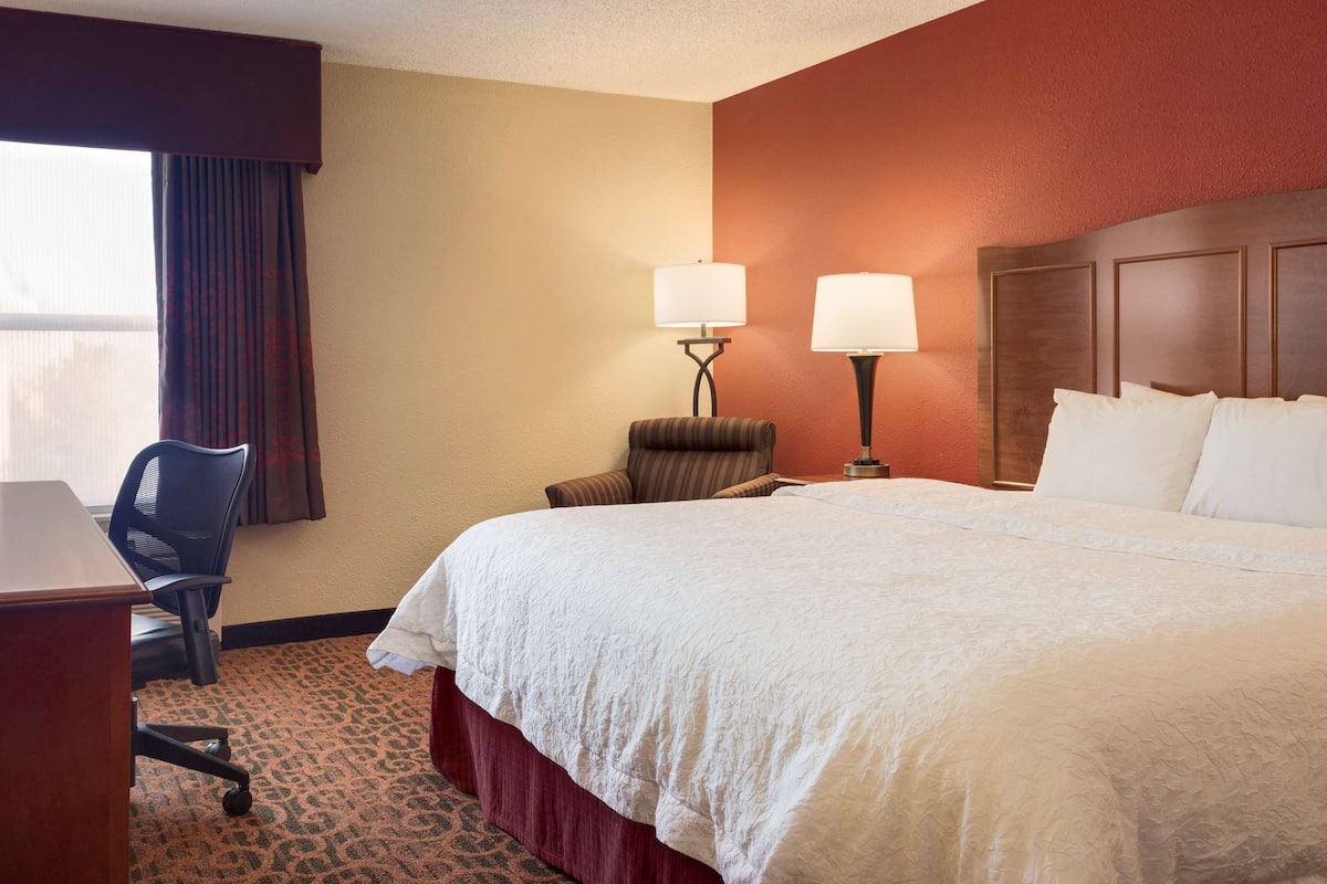 A hotel room with a large bed, two bedside lamps, a desk with a chair, and a window with curtains.