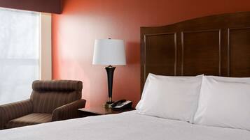 A hotel room with a bed, a chair, a nightstand with a lamp, and a phone.