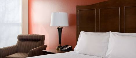 A hotel room with a bed, a chair, a nightstand with a lamp, and a phone.
