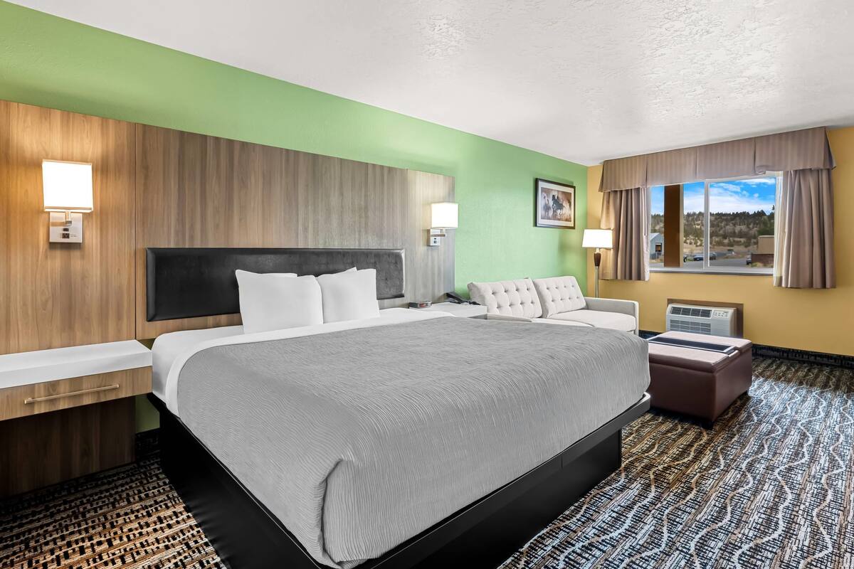 A hotel room with a large bed, a desk, a chair, a window with curtains, and a wall-mounted lamp.