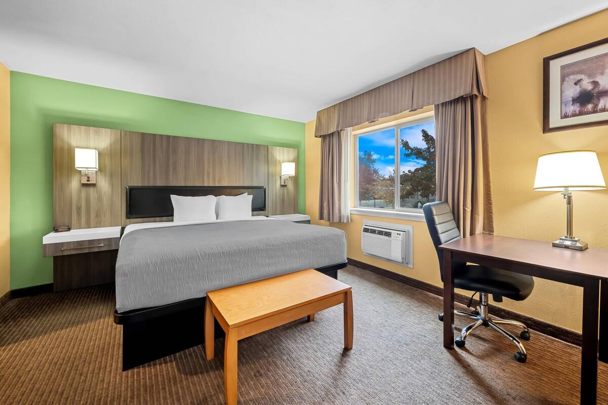 A hotel room with a bed, bedside table, desk, chair, and a window with curtains.