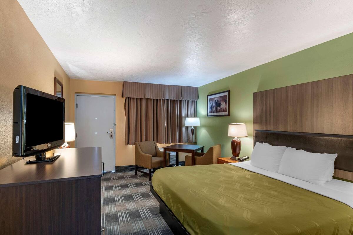 A hotel room with a bed, television, desk, chair, and a painting on the wall.