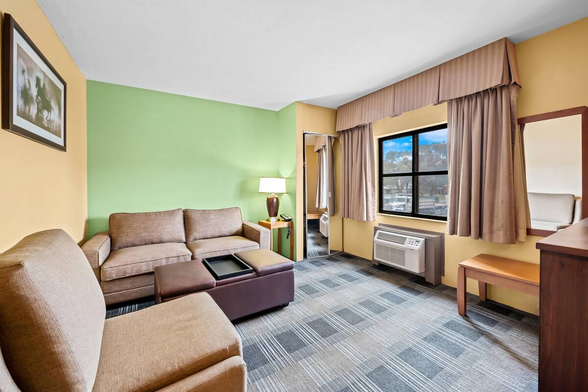 A hotel room with a sofa, a small table, a lamp, a window with curtains, and a radiator.